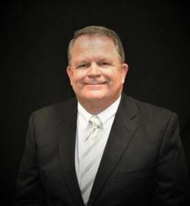 Portrait of Coach Stan Leech in a black suit and striped tie, smiling confidently against a black backdrop.