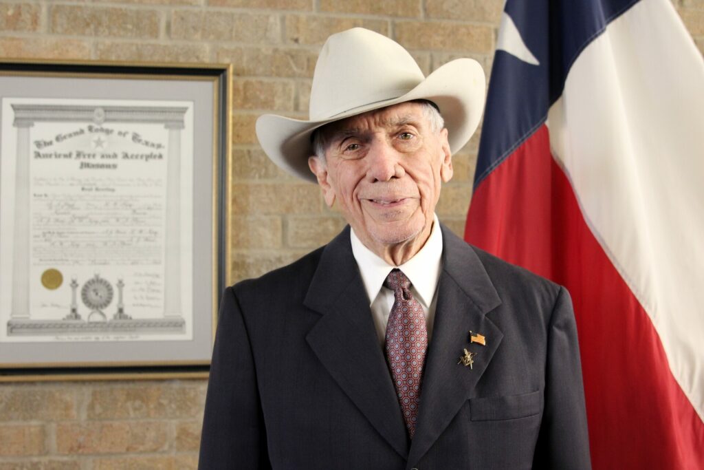 the Kendall Gentleman, Ed Heath, in a Cowboy Hat and Suit Stands Before a Framed Masonic Certificate and the Texas Flag, Embodying Tradition, Leadership, and Service.