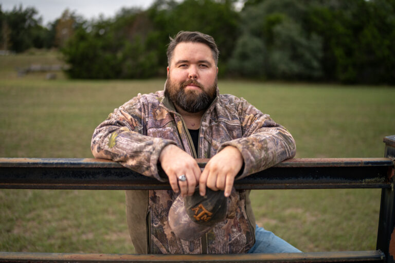 Michael G. Ethridge – Tradition and Leadership in the Texas Hill Country - the Kendall Gentleman Michael Ethridge, the Bearded Publisher of the Kendall Gentleman in a Camouflage Jacket Leans Against a Ranch Gate, Holding a Texas A&m Cap, with the Texas Hill Country Landscape in the Background.