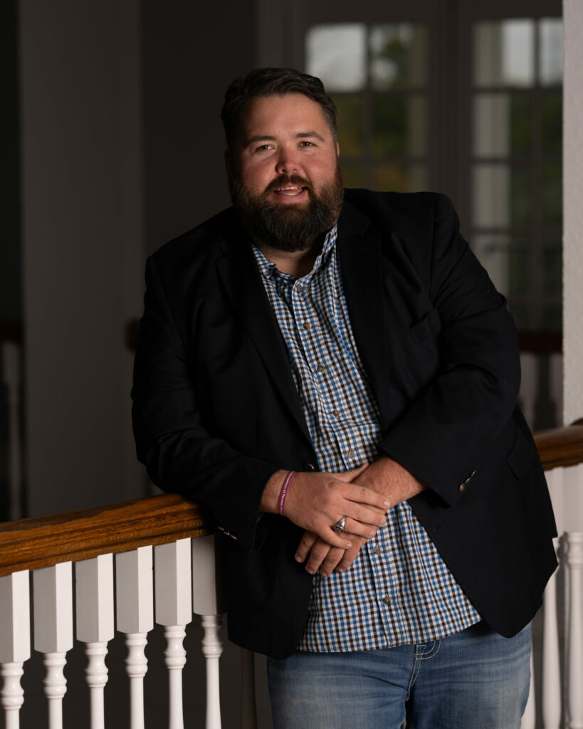 Michael Ethridge – Publisher of the Kendall Gentleman - the Kendall Gentleman Michael Ethridge, Publisher of the Kendall Gentleman, Leaning on a Wooden Railing in a Blue Blazer and Checkered Shirt, Representing Modern Masculinity, Tradition, and Craftsmanship.
