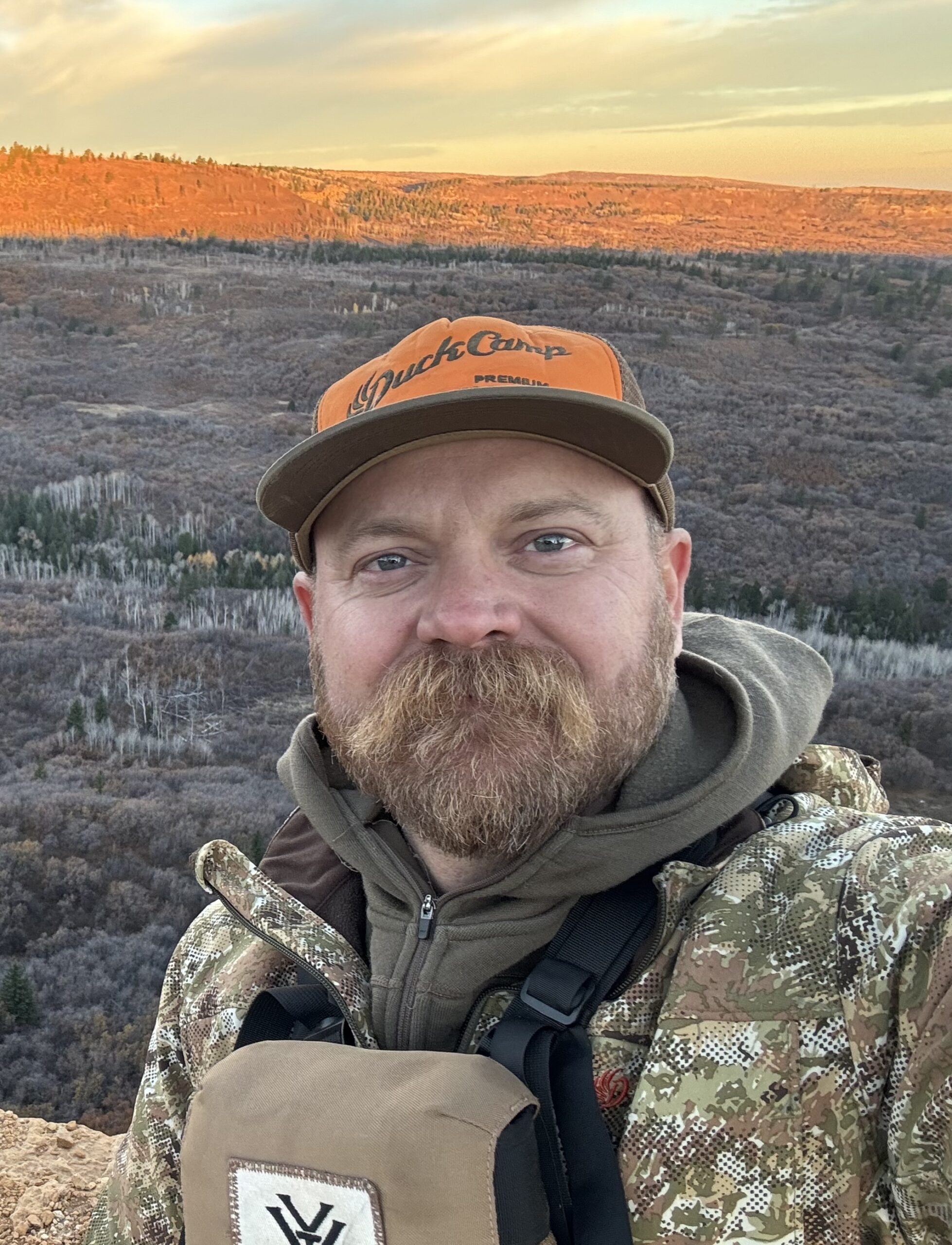 Jed Mazour, Outdoorsman and Contributor to the Kendall Gentleman, Dressed in Camouflage Gear with a Scenic Autumn Mountain Backdrop.