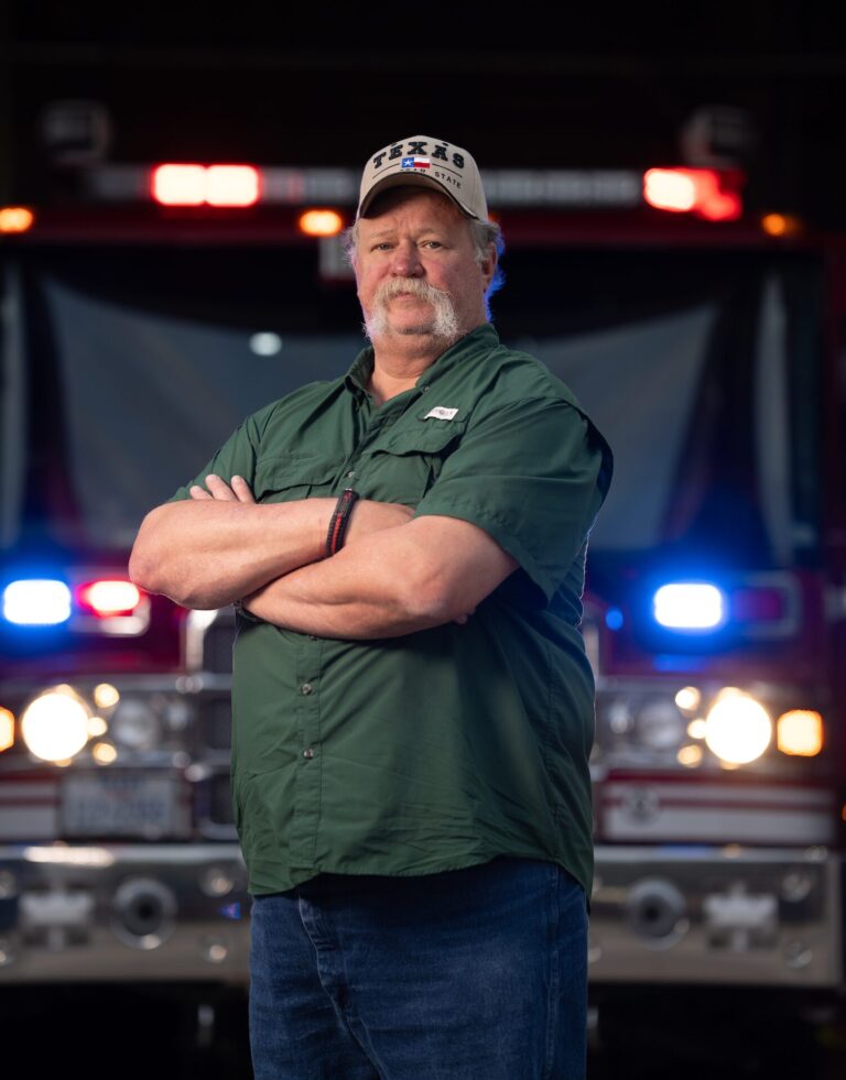 Collins Martin Standing with Arms Crossed in Front of a Firetruck, Wearing a Green Shirt and Texas Cap, Photographed for the Kendall Gentleman.