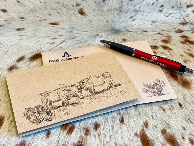 a Cowboy-themed Notecard Set with Longhorn Cattle and Cactus Illustrations, Accompanied by a Texas A&m Real Estate Pen and an Envelope Labeled Frank Addington Jr., Displayed on a Cowhide Surface.