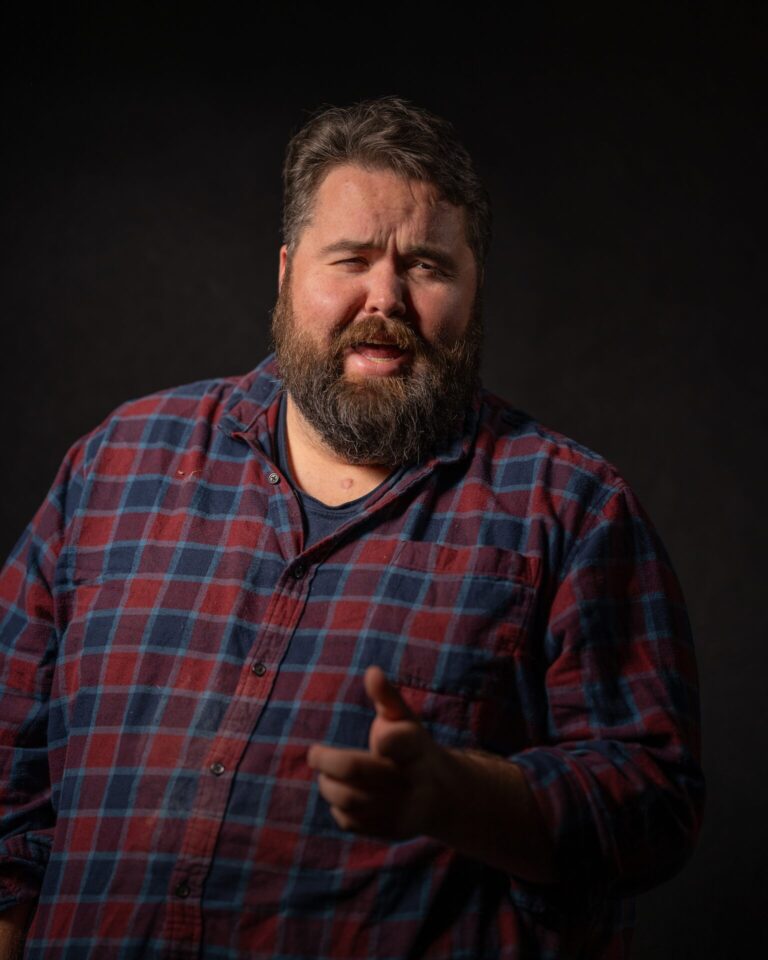 Michael G. Ethridge, Publisher of the Kendall Gentleman, in a Red and Blue Flannel Shirt, Pictured Against a Dark Background with Dramatic Studio Lighting.