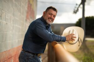 Cody Davenport leans casually on a fence, smiling with a cowboy hat in hand and dressed in denim.