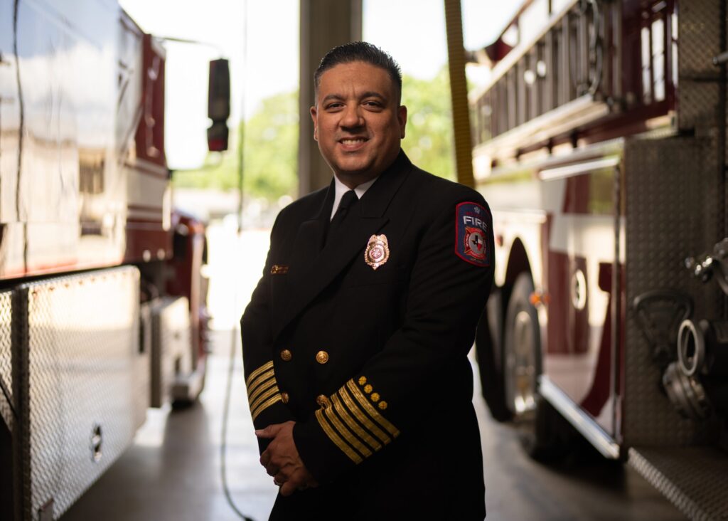 Fire Chief Manny Cazares Stands Between Fire Trucks in Uniform, Smiling with Pride and Professionalism.