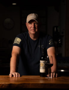 “Andy Ivankovich at Singing Water Vineyards in Comfort, Texas, with a bottle of Freedom red wine and a glass on the tasting room bar.”