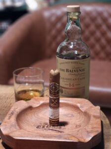 A Montecristo Espada Ricasso cigar stands in a wooden ashtray next to a glass and bottle of Balvenie 14 Year Caribbean Cask Scotch.