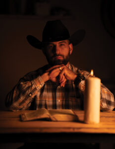 Casey Barker in a cowboy hat, seated at a table with an open Bible and candle, hands clasped in quiet reflection.