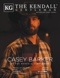 Casey Barker holds a Bible close to his chest, wearing a cowboy hat and plaid shirt, featured on the September 2025 cover of The Kendall Gentleman.