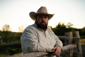Michael G. Ethridge stands by a wooden fence at sunset, wearing a cowboy hat and patterned shirt, looking directly into the camera with a serious expression.
