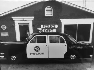 Black-and-white photo of a vintage Police Department cruiser parked outside 114 E Texas, with classic signage and building facade.
