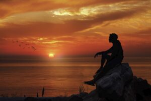 Silhouette of a man sitting on a rock overlooking the ocean at sunset, deep in thought as birds fly across a vivid orange sky.
