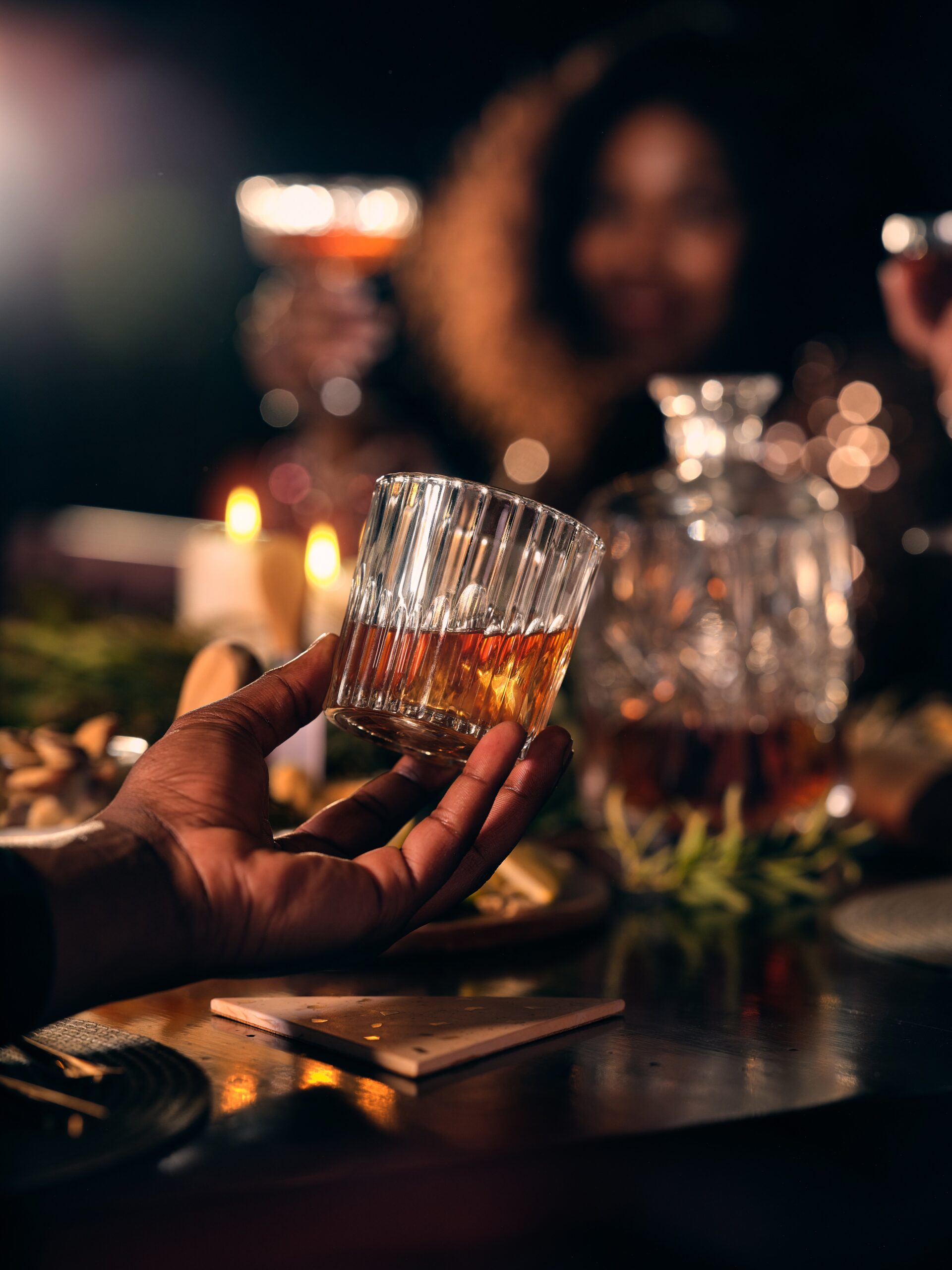 Close-up of a Hand Holding a Glass of Whiskey at a Warmly Lit Table, with Friends and a Crystal Decanter in the Blurred Background.