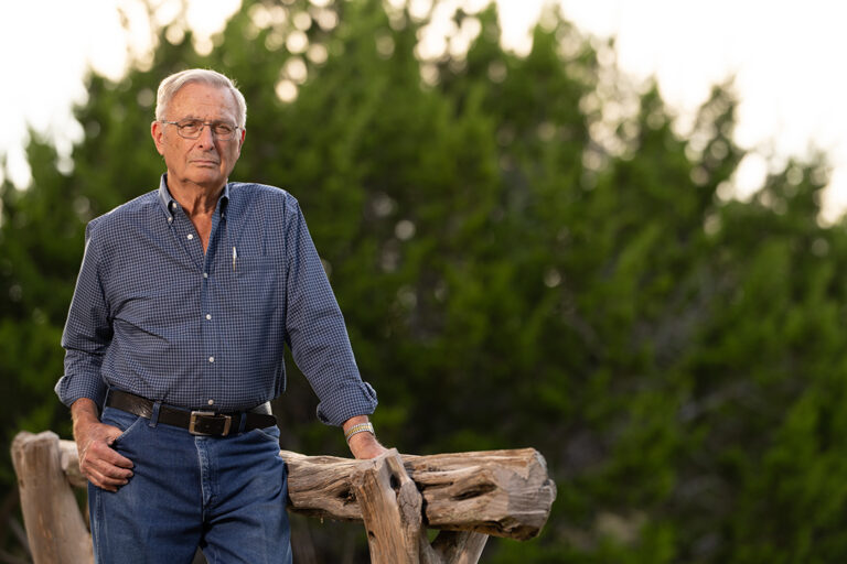 Ken Nietenhoefer stands outdoors by a wooden fence, wearing a blue checkered shirt and glasses, featured in The Kendall Gentleman cover story.