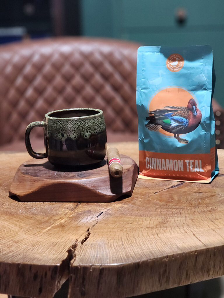 Coffee mug, Romeo y Julieta 1875 Bully cigar, and Dirty Duck Cinnamon Teal coffee on a rustic wooden table.