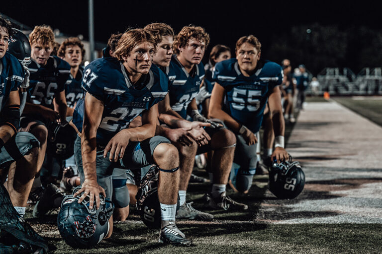 High school football players kneeling on the sideline during a night game, wearing blue uniforms and holding helmets.