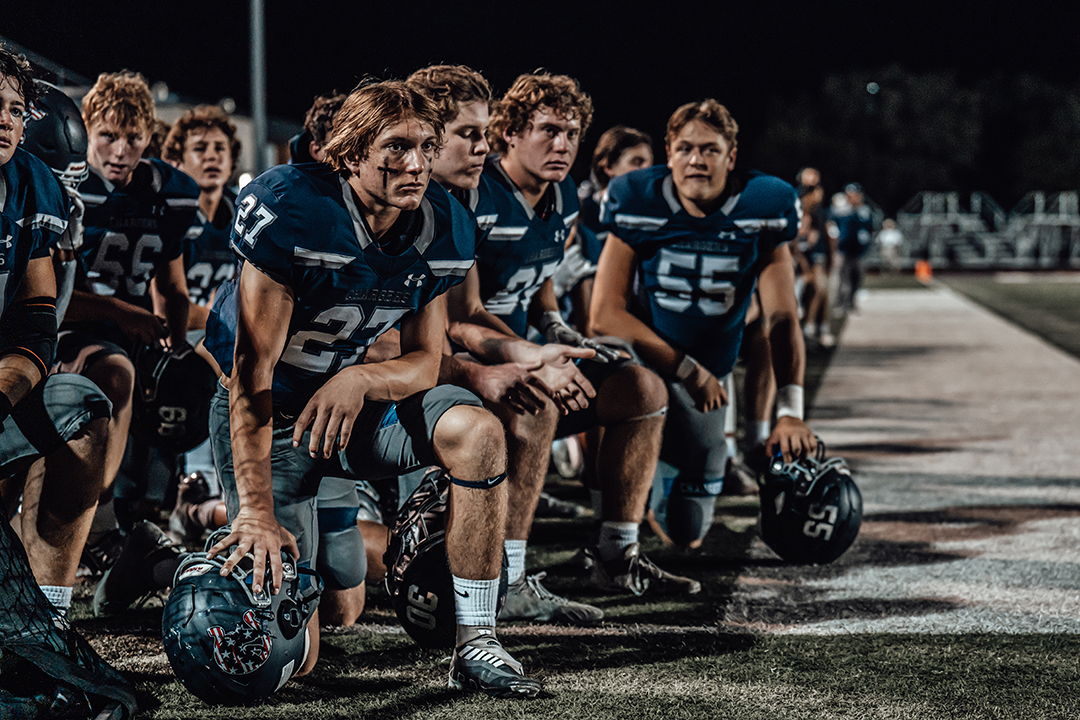 High School Football Players Kneeling on the Sideline During a Night Game, Wearing Blue Uniforms and Holding Helmets.