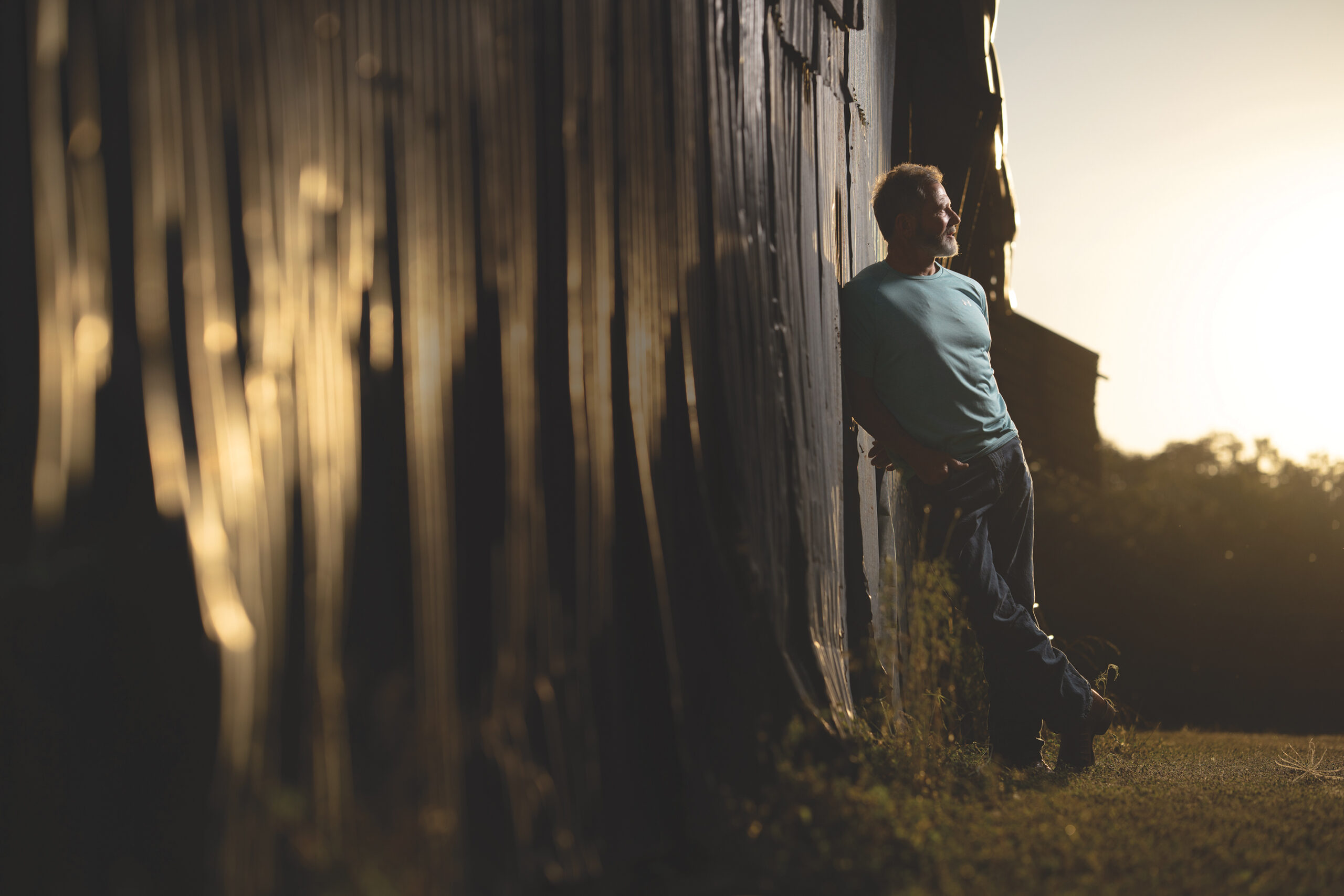 Donny May Leans Against a Barn Wall at Sunset on His Bergheim Ranch, Reflecting Strength, Humility, and Faith.