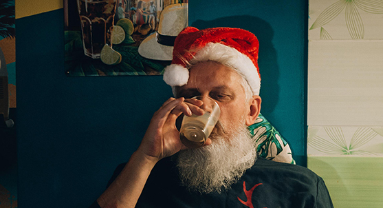 Older Man Wearing a Santa Hat Drinks from a Glass, Capturing the Humorous Spirit of Small-town Christmas Memories.
