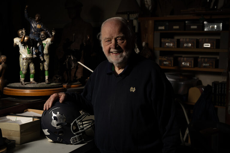 Elderly Sculptor Smiling in His Studio Beside a Detailed Football Sculpture and a Championship Football Helmet.