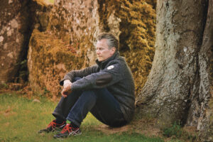 Man sitting thoughtfully against a tree in a natural outdoor setting with rock formations in the background.