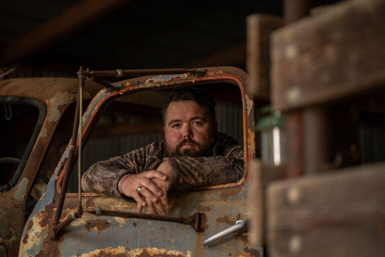 Bearded Man Leaning out of the Window of a Weathered Vintage Truck Inside a Rustic Barn.