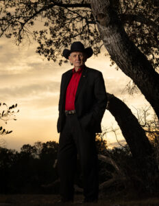 General Leroy Sisco standing outdoors at sunset wearing a black cowboy hat and red shirt, representing a lifetime of military service and leadership.