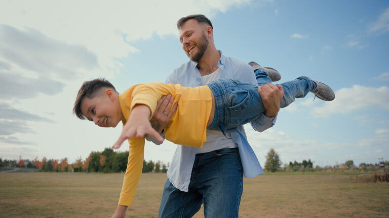 Father Holding His Son Airplane-style While Playing in a Field, Showing Joy, Connection, and the Importance of Being Present with Family