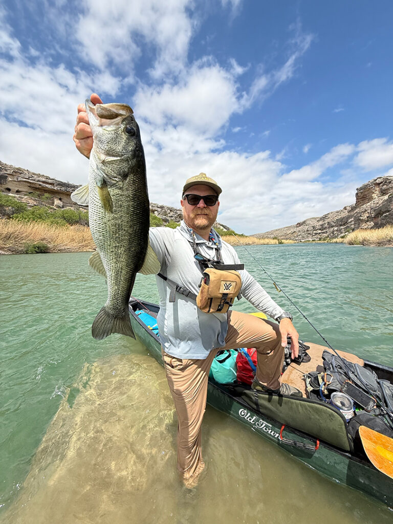 Man in a Wide-brimmed Hat and Sunglasses Holds Up a Large Bass Beside a Kayak in a Shallow, Green River Canyon Scene.