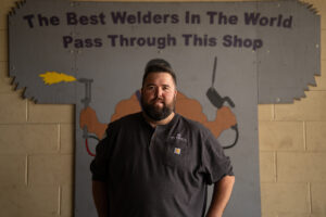 Michael G. Ethridge standing inside a welding shop beneath a sign reading “The Best Welders In The World Pass Through This Shop,” reflecting craftsmanship, discipline, and leadership.