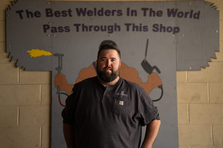 Michael G. Ethridge Standing Inside a Welding Shop Beneath a Sign Reading “the Best Welders in the World Pass Through This Shop,” Reflecting Craftsmanship, Discipline, and Leadership.