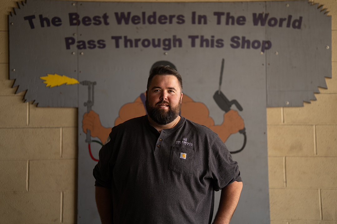 Michael G. Ethridge Standing Inside a Welding Shop Beneath a Sign Reading “the Best Welders in the World Pass Through This Shop,” Reflecting Craftsmanship, Discipline, and Leadership.