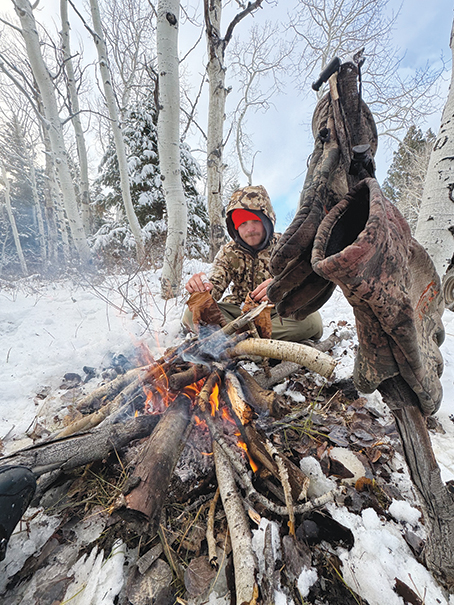 Hunter Tending a Campfire Beside Hanging Gear in a Snowy Mountain Forest