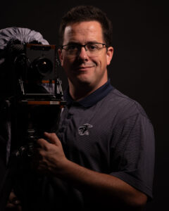 Portrait of a Man Wearing Glasses Beside a Large-format View Camera on a Dark Backdrop.