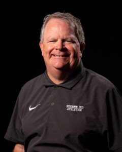 Smiling Man in a Black Polo with Boerne Isd Athletics Logo Against a Black Background.