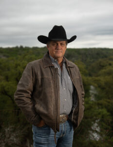 Glynn Praesel in a black cowboy hat and brown leather jacket standing in front of wooded Texas Hill Country