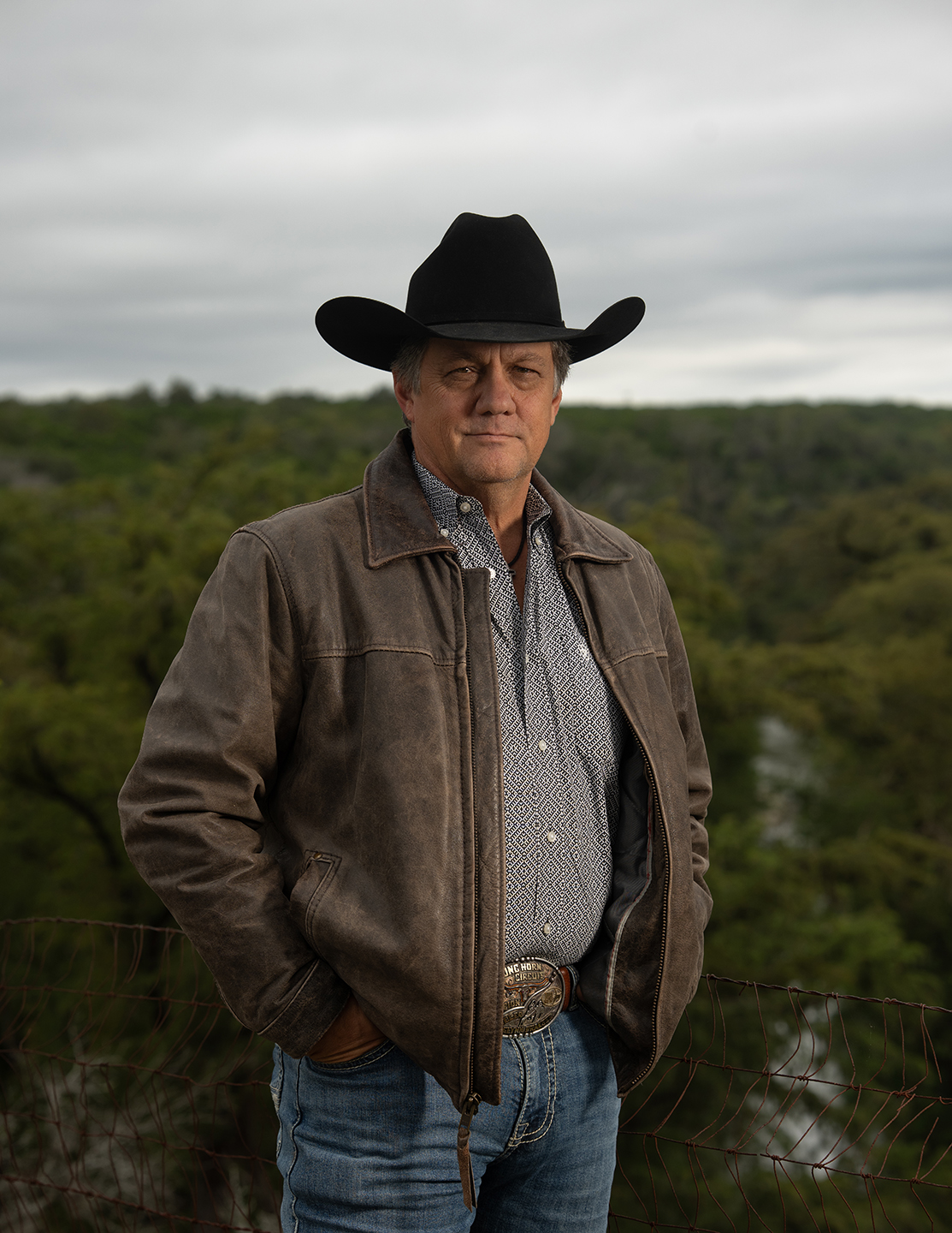 Glynn Praesel in a Black Cowboy Hat and Brown Leather Jacket Standing in Front of Wooded Texas Hill Country