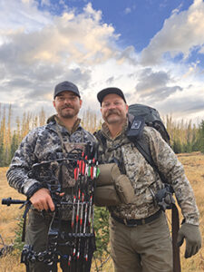 Two bowhunters in camouflage with compound bows and packs standing in a high-country meadow