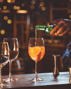 A hand pouring from a green bottle into a stemmed glass holding an orange cocktail with ice and an orange slice
