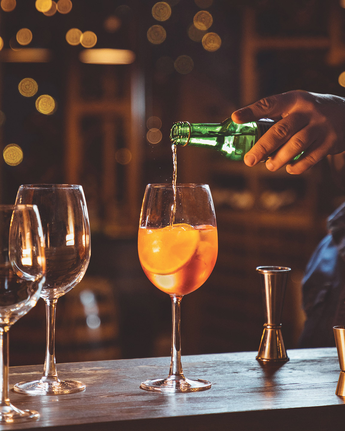 a Hand Pouring from a Green Bottle into a Stemmed Glass Holding an Orange Cocktail with Ice and an Orange Slice